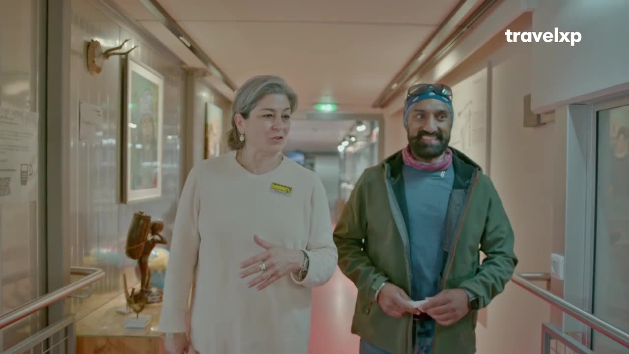 A woman in a cream sweater gestures as she walks down a hallway, a man in a green jacket following close behind. They are exploring a space, perhaps a museum or gallery, with artwork displayed on the walls.