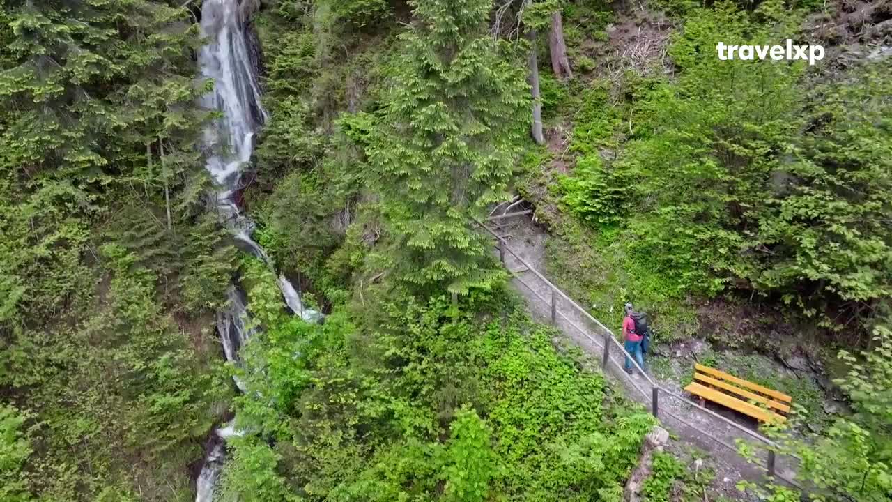 A lone hiker walks up a winding path beside a cascading waterfall. Lush green trees and foliage surround the trail, with a wooden bench offering a place to rest.