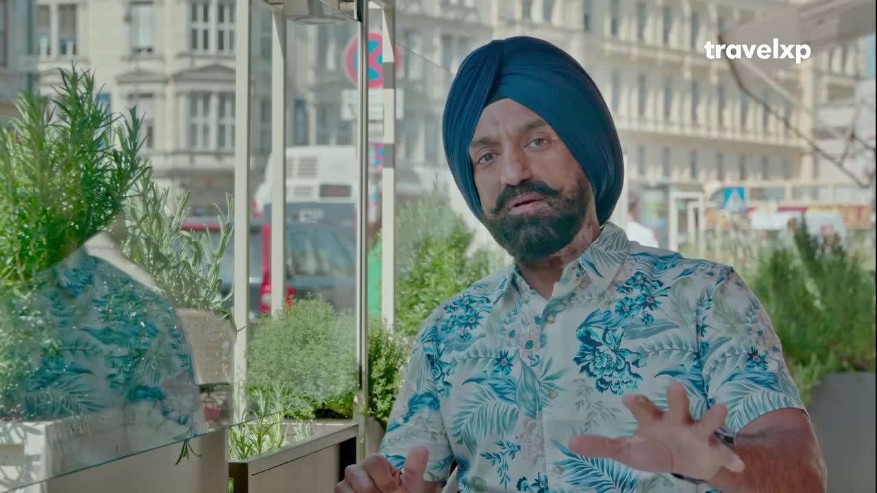 A man in a blue turban and a floral shirt gestures with his hands. Behind him, a bus passes by a building with ornate architecture.
