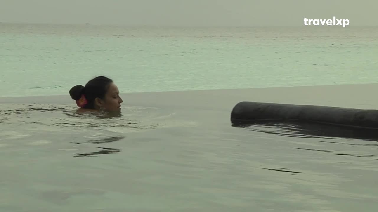 A woman swims in an infinity pool, her hair pulled back in a bun. The calm water stretches out to meet the horizon, reflecting the overcast sky.