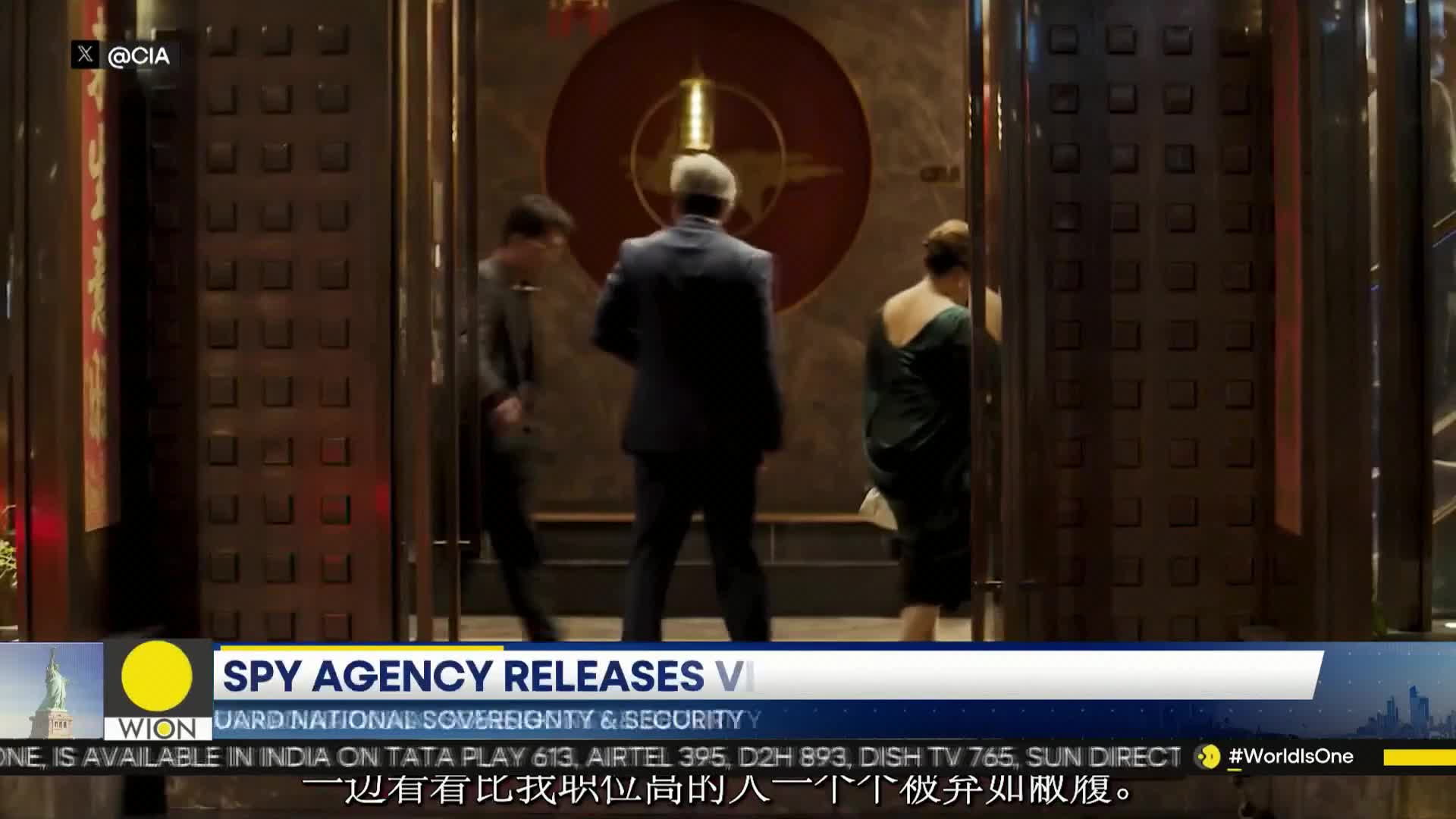 A man in a dark suit walks past a large circular emblem. A woman in a green dress follows him, her back to me. Another person, also in dark clothing, moves quickly to their left.