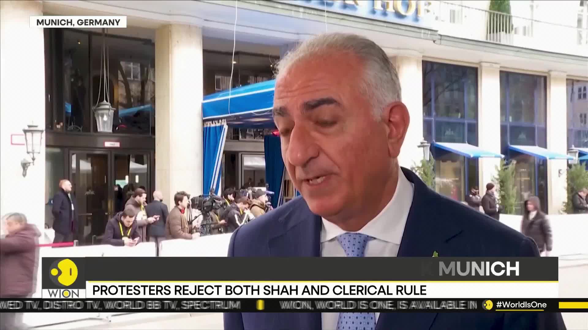 A man in a suit speaks into a microphone, a crowd of people and news cameras gathered behind him. The backdrop is a building with large windows and blue awnings.