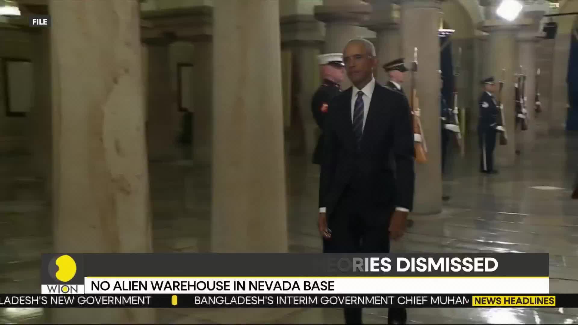 A man in a dark suit walks past, his gaze forward. Behind him, guards in uniform stand at attention with rifles.