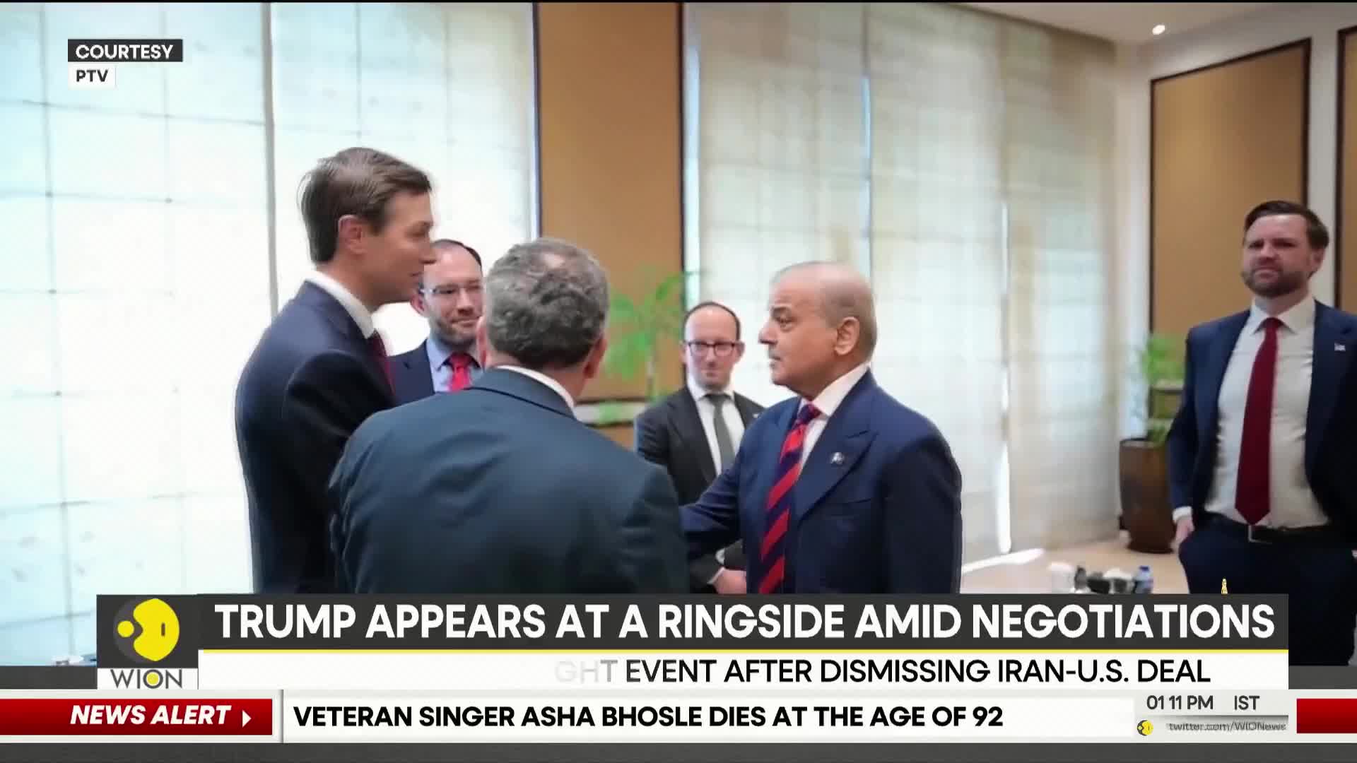 A group of men in suits stand in a room, some engaged in conversation. A news ticker at the bottom of the screen announces the passing of veteran singer Asha Bhosle.