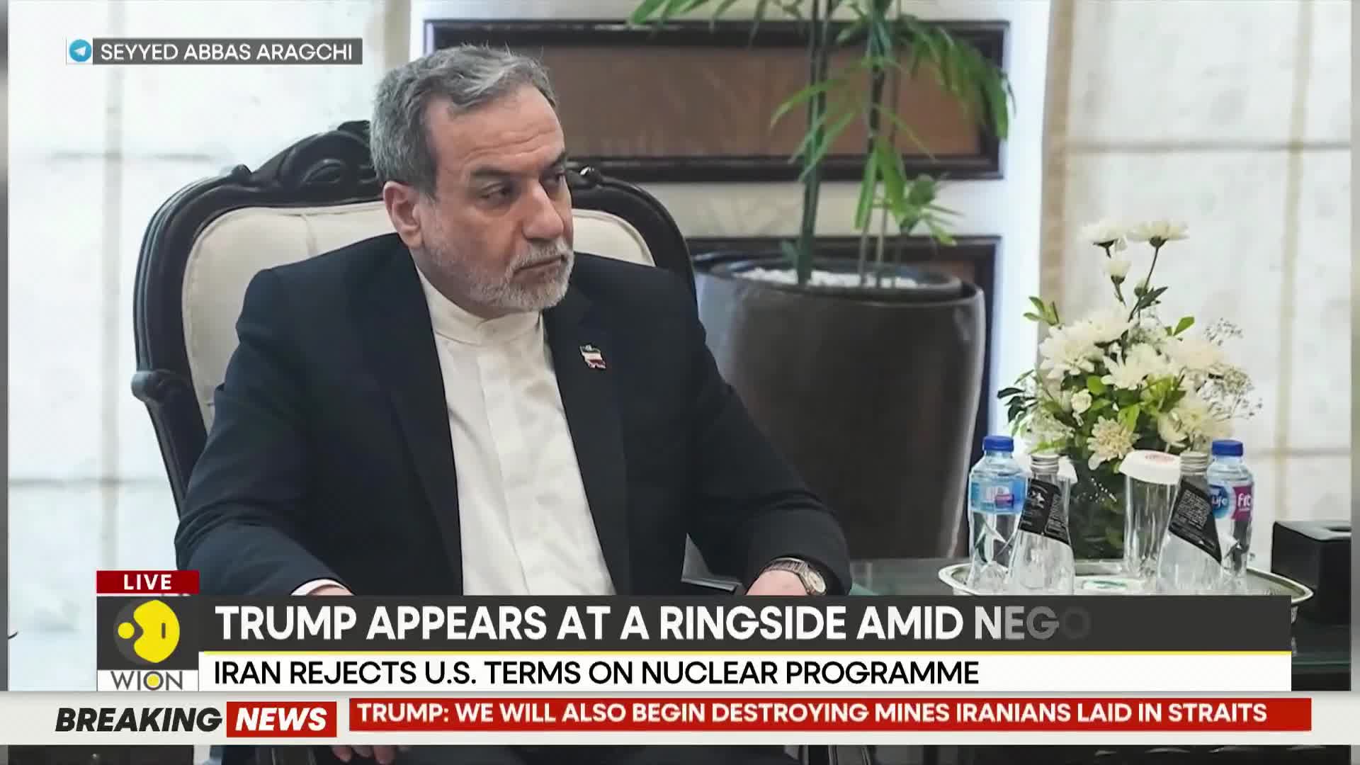 A man in a suit sits in a plush chair, his gaze fixed on something just beyond the frame. A bouquet of white flowers and several water bottles rest on a table before him, while a chyron from WION reports on Iran's rejection of US terms.