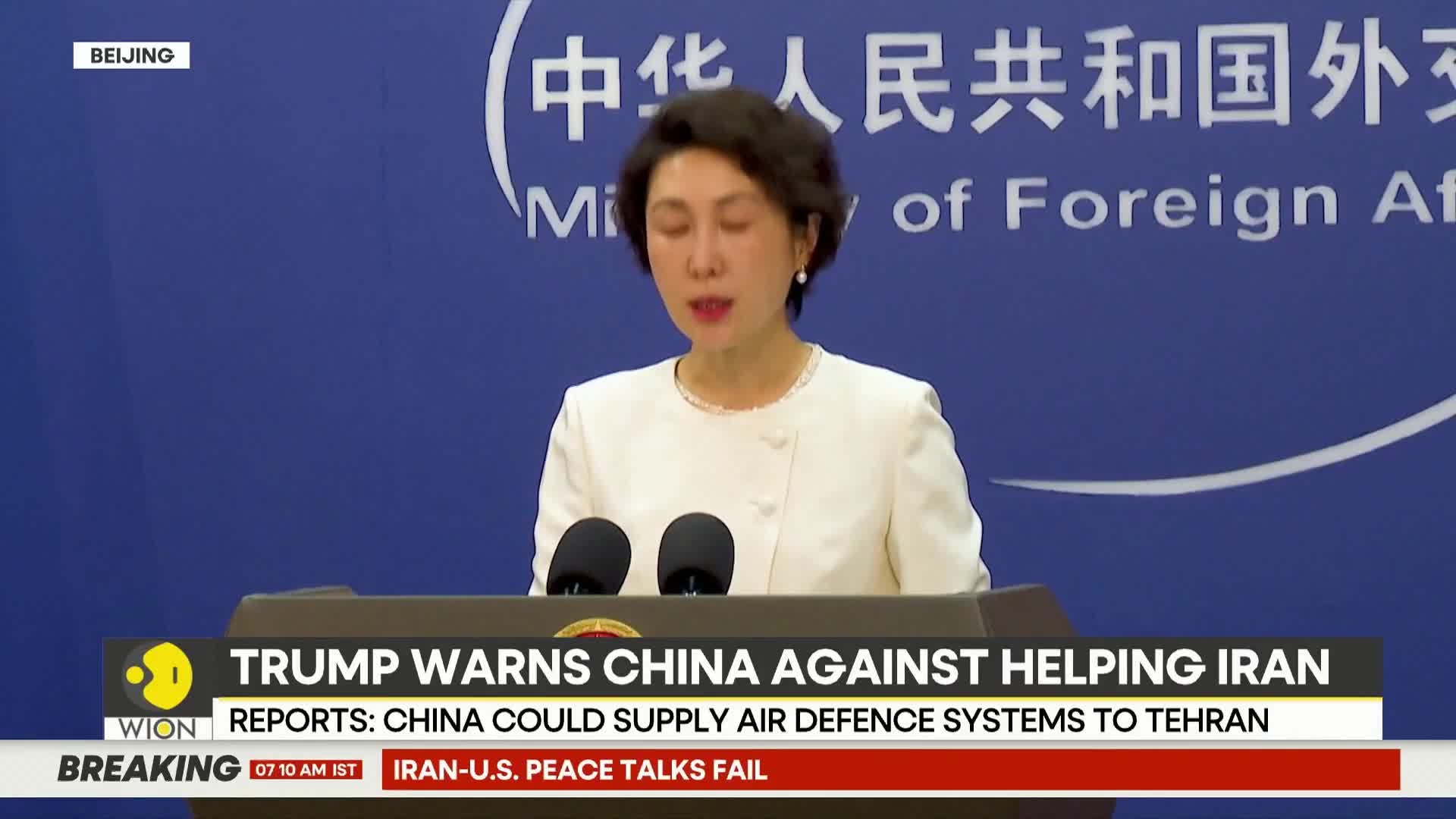 A woman stands at a podium, speaking into two microphones. A news ticker at the bottom of the screen reports on international relations.