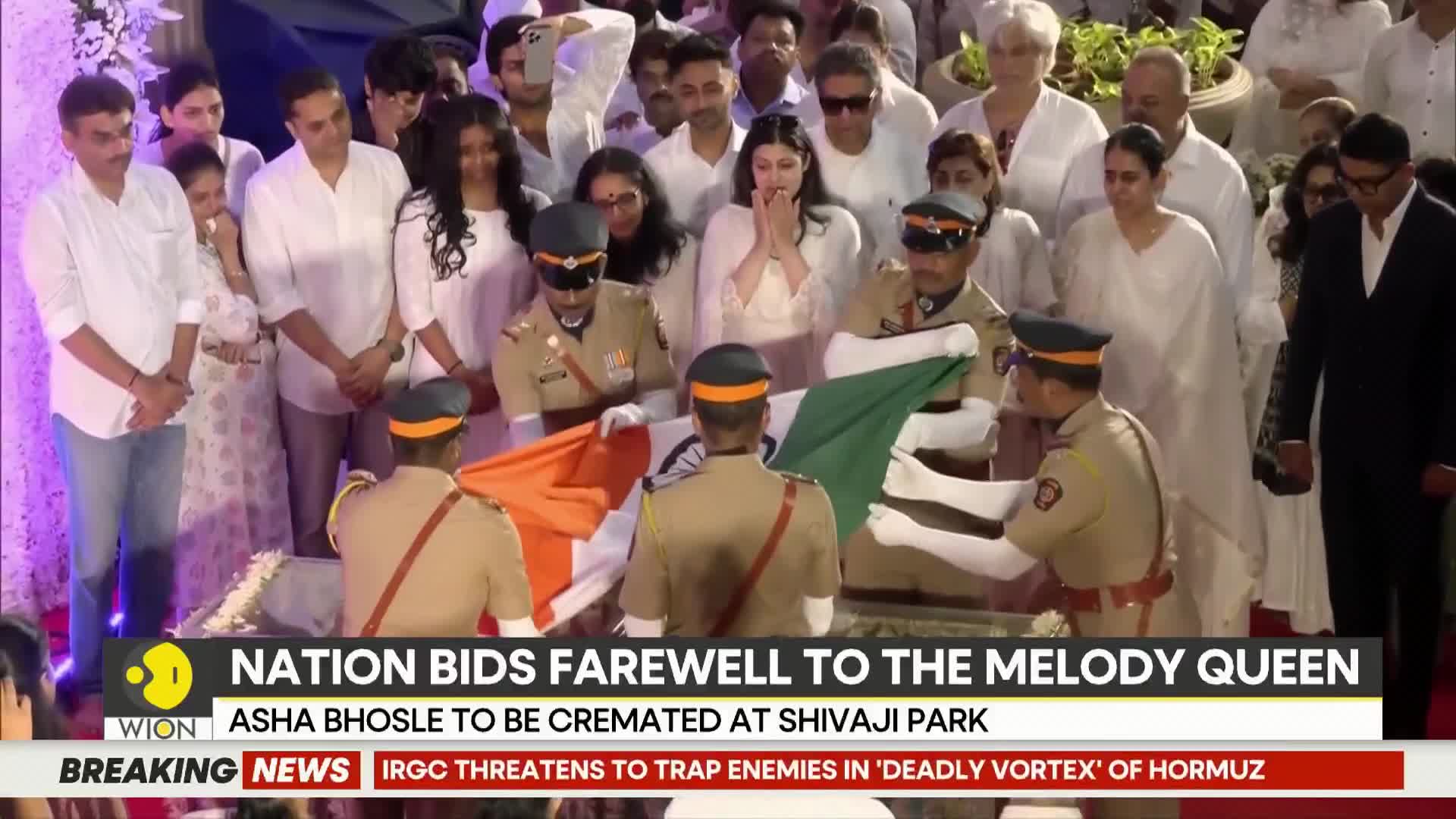 Uniformed officers carefully fold the Indian flag over a casket. A crowd of mourners, many in white, stands somberly behind them.