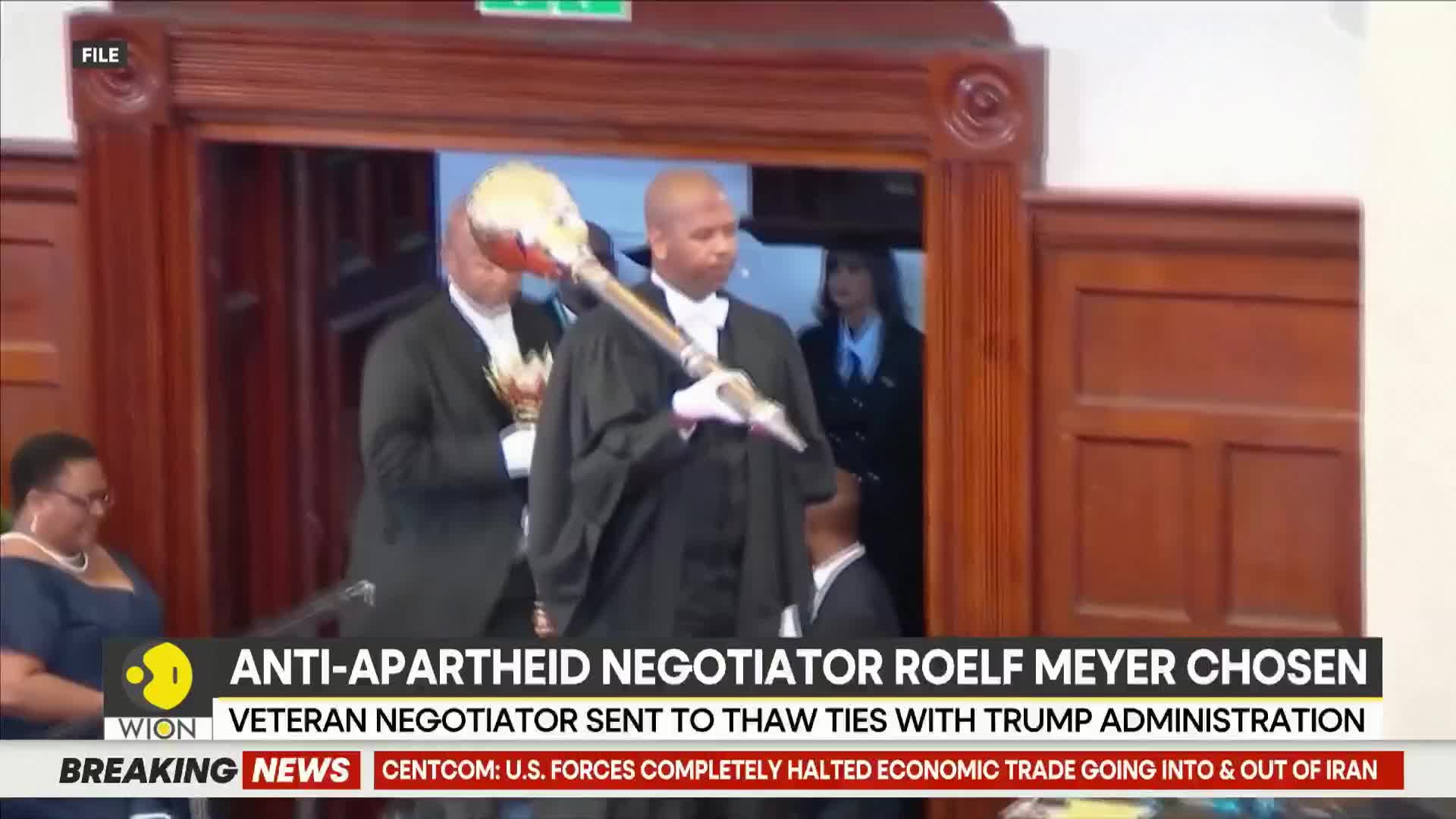 A man in a black robe carries a ceremonial mace, its head adorned with white and red feathers. Behind him, others in formal attire stand in a grand, wood-paneled room.