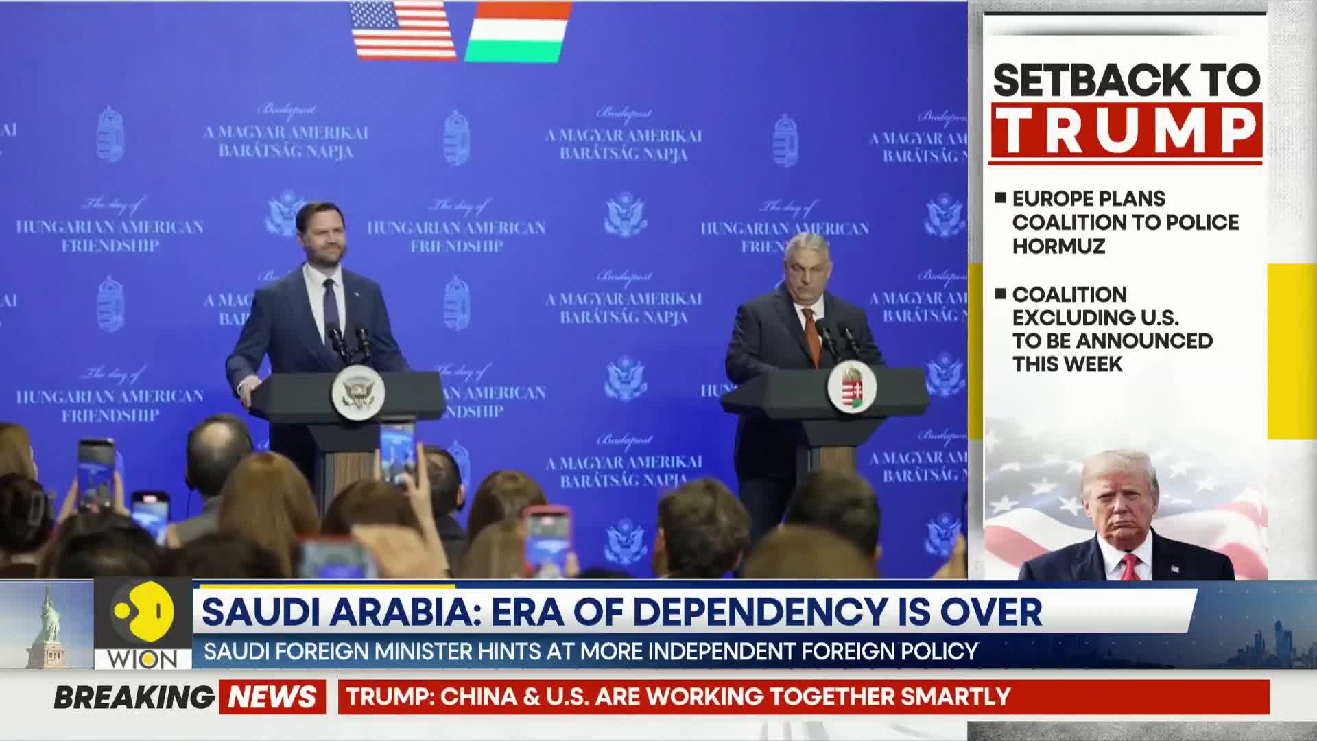 Two men stand at podiums in front of a blue backdrop, speaking to a crowd holding up phones. A news ticker at the bottom displays "SAUDI ARABIA: ERA OF DEPENDENCY IS OVER" and "BREAKING NEWS TRUMP: CHINA & U.S. ARE WORKING TOGETHER SMARTLY."