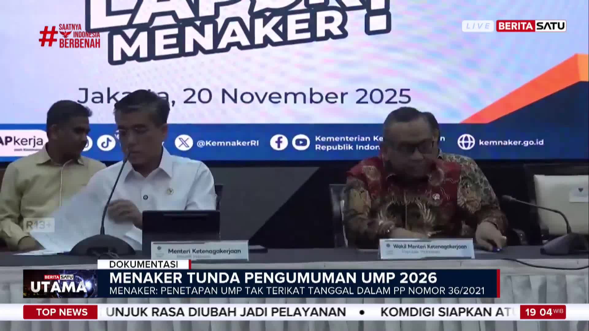 Three men sit at a table, one in a white shirt is holding papers, while the man to his right adjusts his glasses. The BeritaSatu English broadcast is reporting on a Menaker announcement regarding UMP 2026.
