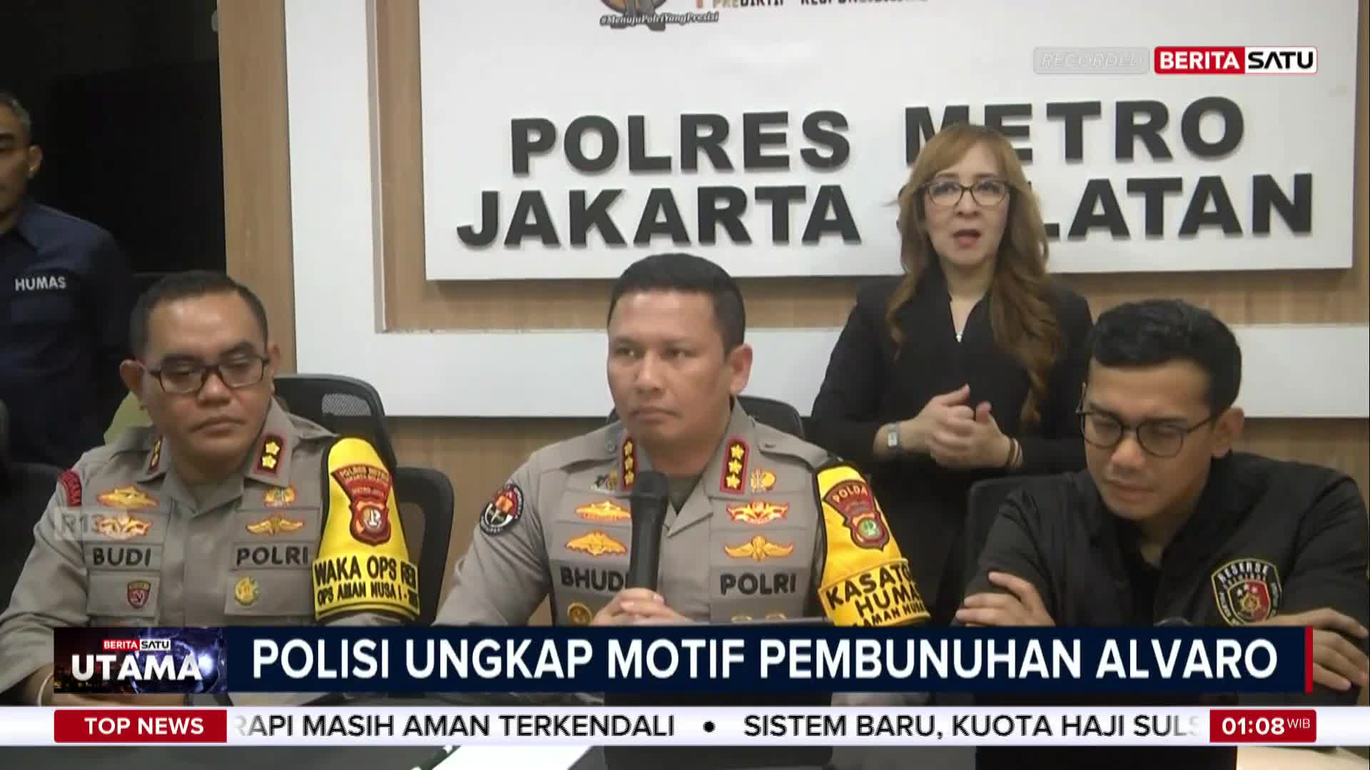 Three uniformed police officers sit at a table, while a woman in a black blazer and glasses speaks behind them. A BeritaSatu English news banner at the bottom announces "POLISI UNGKAP MOTIF PEMBUNUHAN ALVARO."
