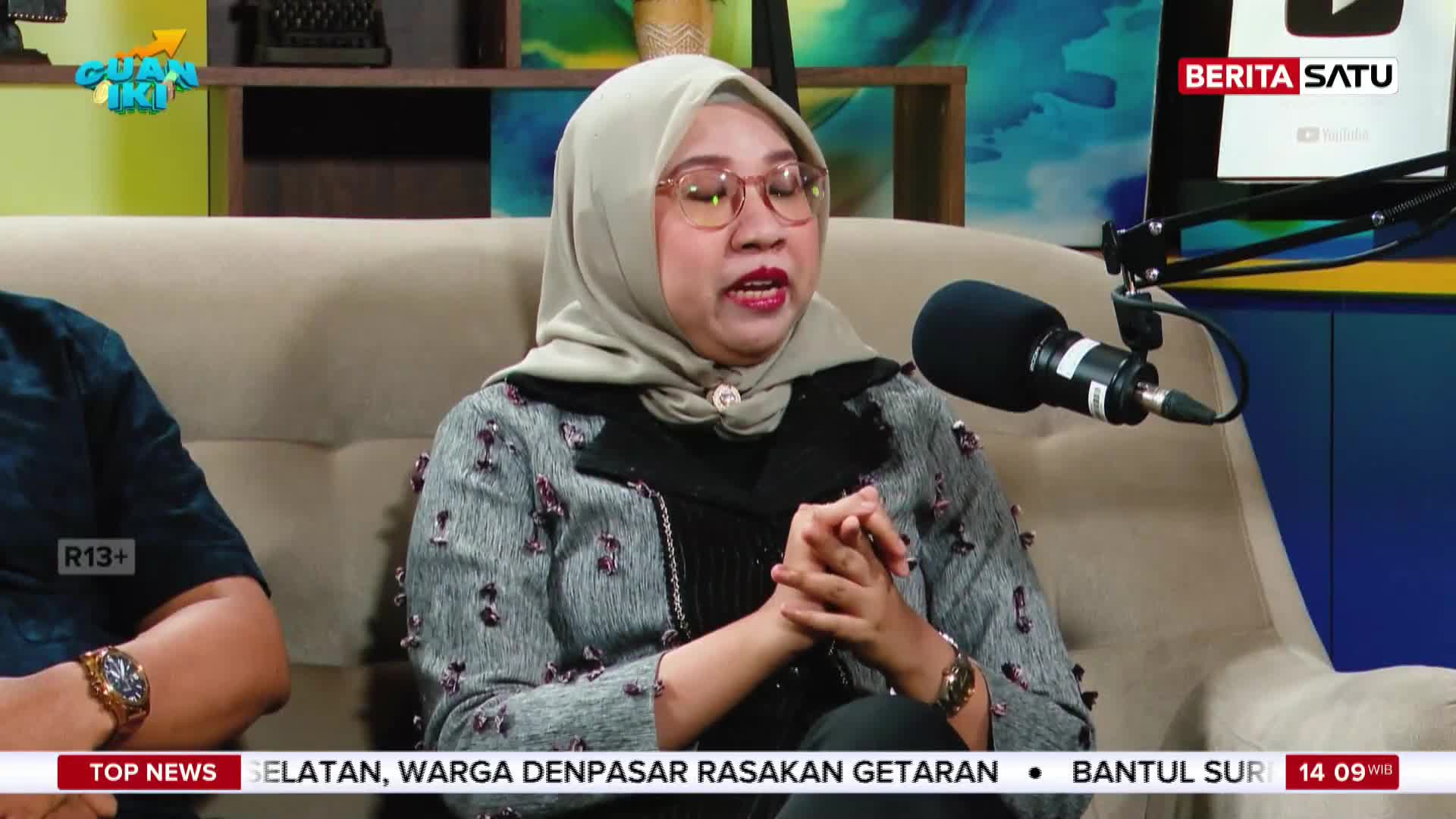 A woman in a hijab speaks into a microphone, her hands clasped. On the screen behind her, Berita Satu's logo flashes. A woman in a hijab speaks into a microphone, her hands clasped. On the screen behind her, Berita Satu's logo flashes.