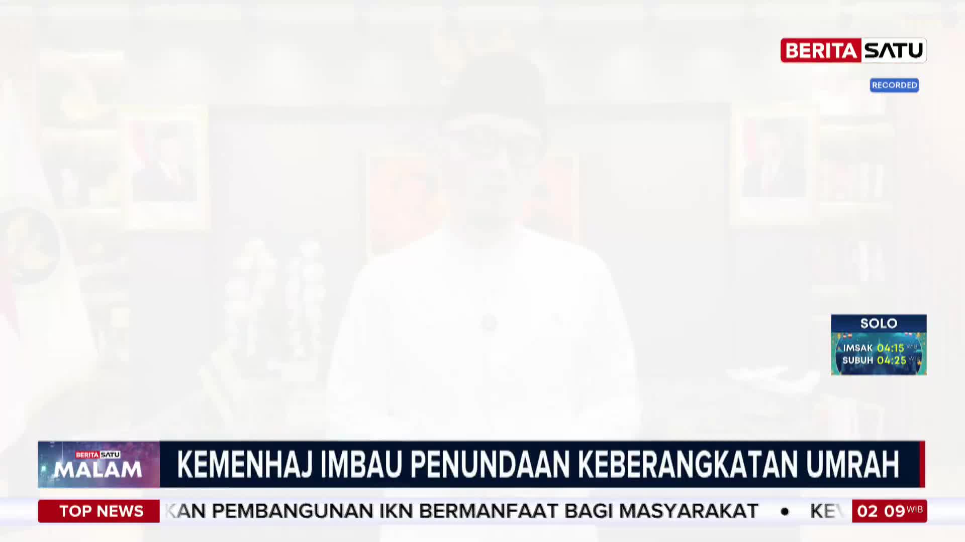 A man in a white shirt and cap stands behind a desk, speaking to the camera. A news ticker scrolls across the bottom of the screen, reporting on the Ministry of Religious Affairs urging a postponement of Umrah departures. A man in a white shirt and cap stands behind a desk, speaking to the camera. A news ticker scrolls across the bottom of the screen, reporting on the Ministry of Religious Affairs urging a postponement of Umrah departures.