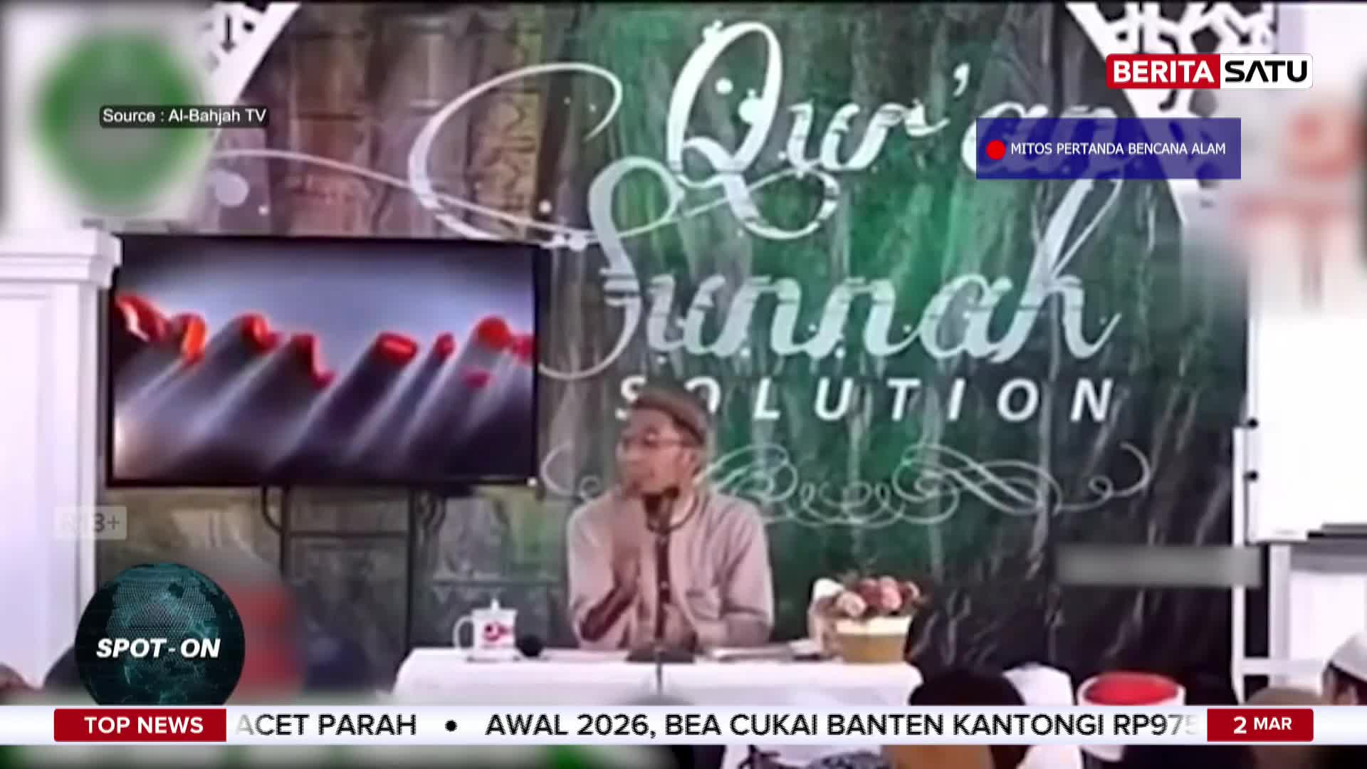 A man in a light brown shirt and cap speaks into a microphone at a table. Behind him, a large green banner displays "Qur'an Sunnah SOLUTION" in white script. A man in a light brown shirt and cap speaks into a microphone at a table. Behind him, a large green banner displays "Qur'an Sunnah SOLUTION" in white script.