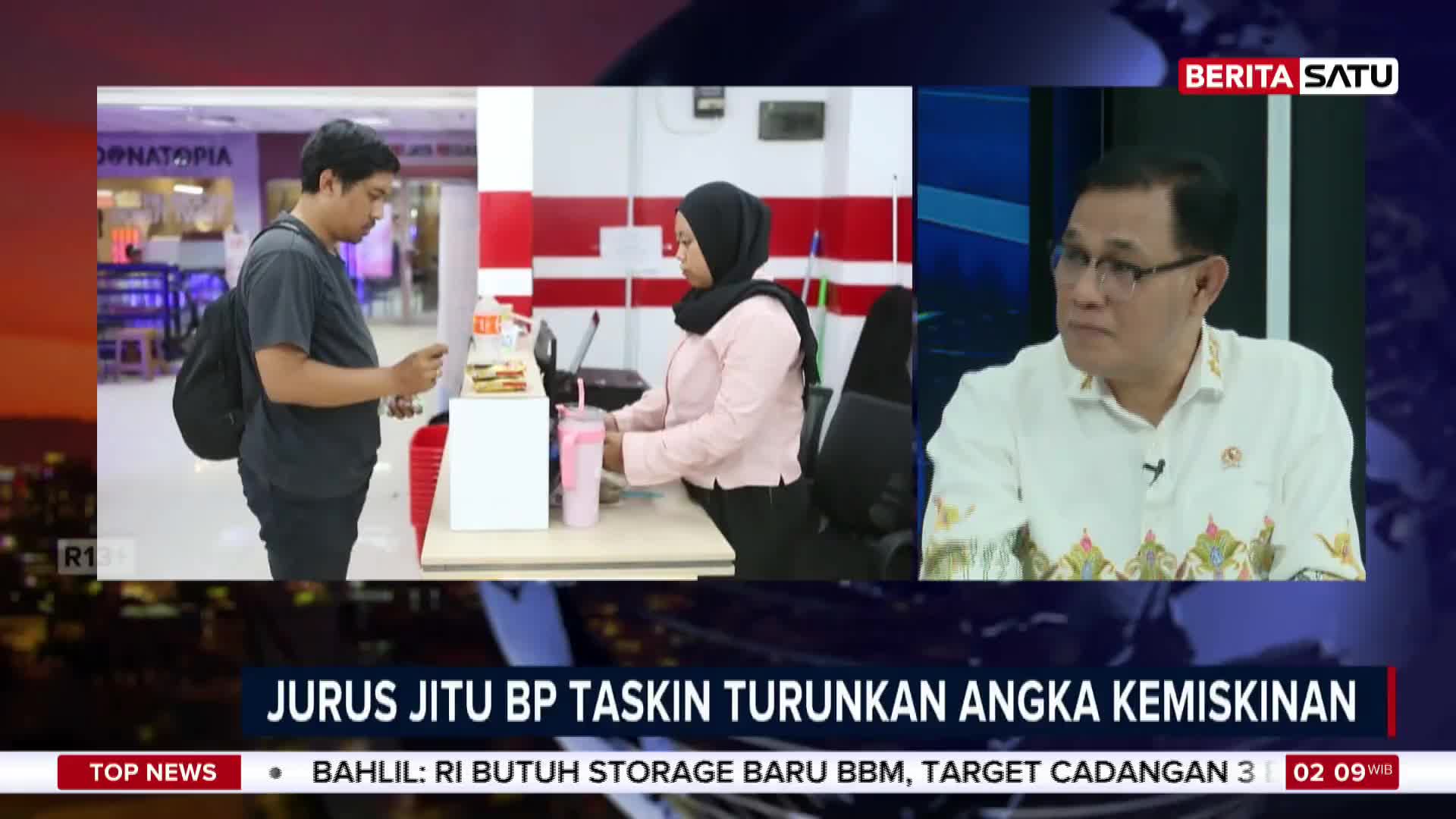 A man in a dark t-shirt and backpack stands at a counter, holding his phone. A woman in a pink shirt and hijab serves him from behind the counter.