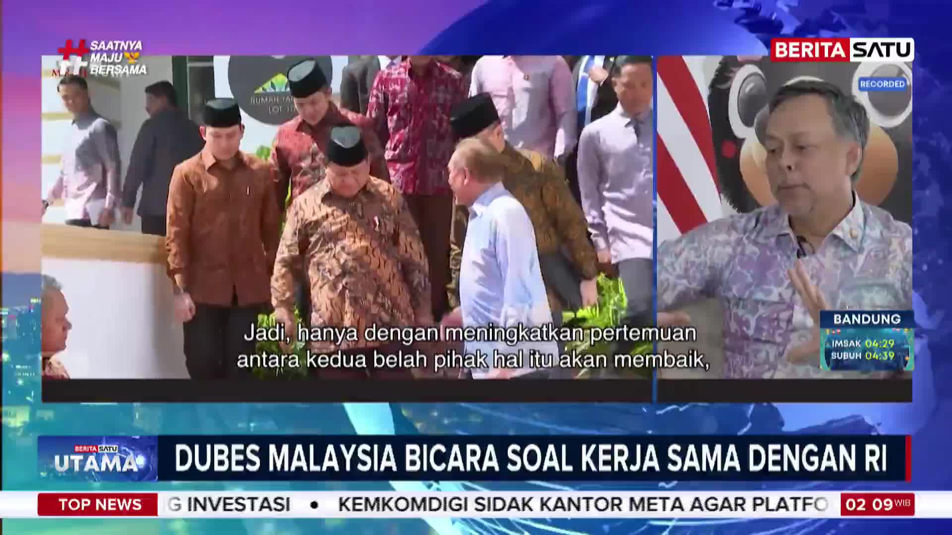 A group of men in traditional Indonesian attire walk past, some wearing peci caps. On the right, a man in a patterned shirt speaks, his hands gesturing as he addresses the camera.