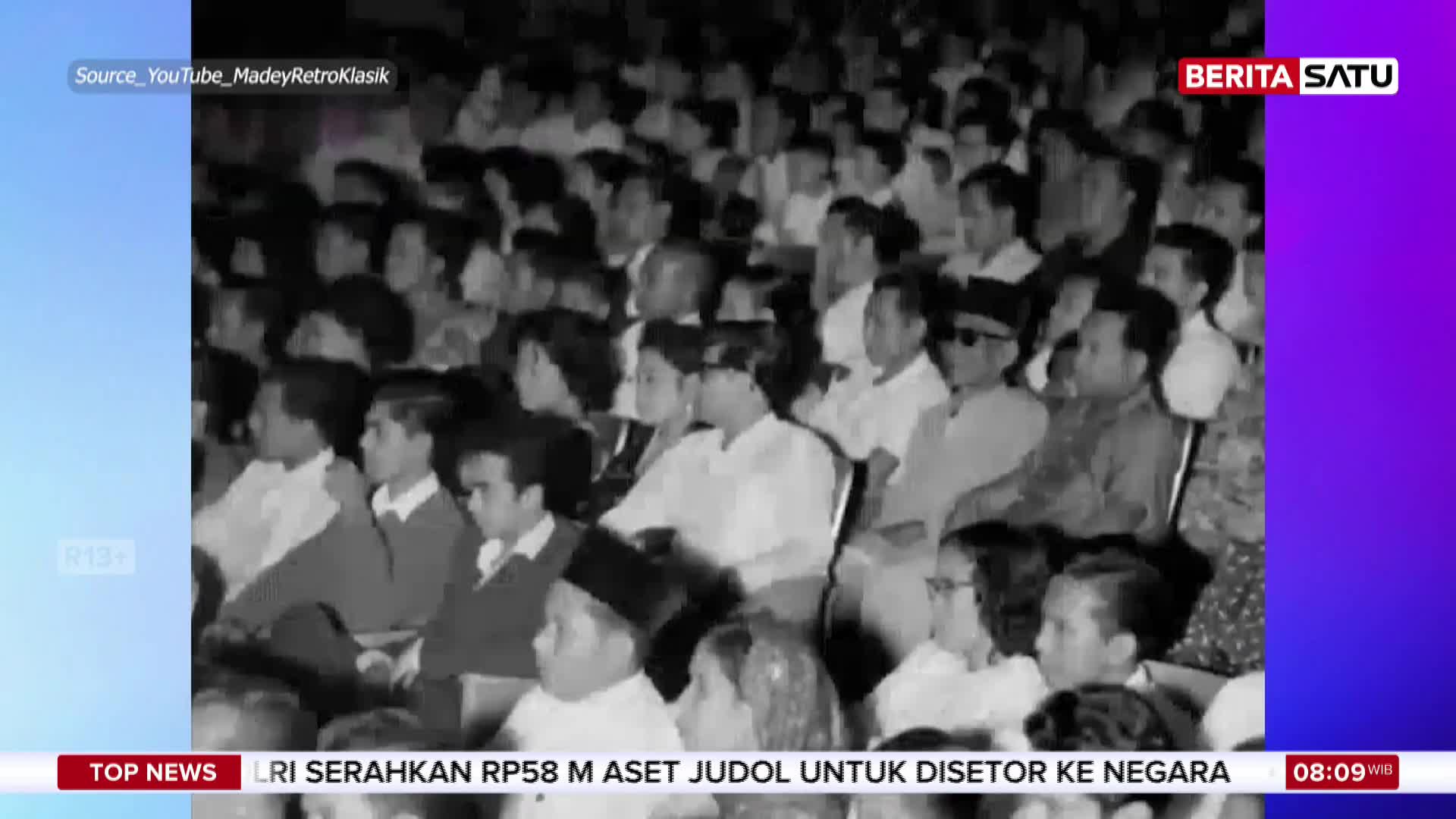 A crowd of people in formal attire fills the seats, their faces turned towards something unseen. A man in a peci hat and sunglasses sits prominently in the foreground, his gaze fixed ahead.