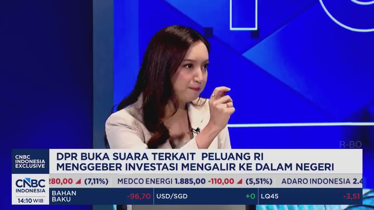 A woman speaks animatedly, gesturing with her fingers. Below her, stock tickers scroll across the screen, displaying Indonesian company names and their performance.