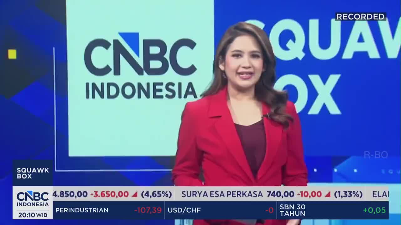 A woman in a red blazer stands before a bright screen displaying the CNBC Indonesia logo and "SQUAWK BOX." Below her, ticker tape scrolls with financial data, including stock prices and currency exchanges.