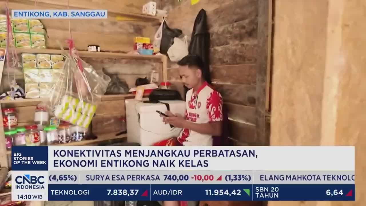 A young man in a red and white shirt sits looking at his phone in a small, cluttered room. Shelves above him are packed with packaged goods and jars.