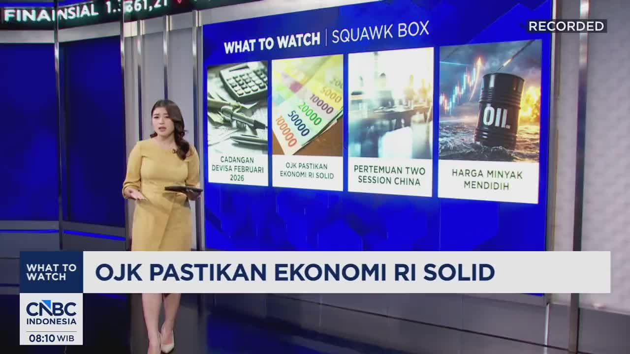 A woman in a yellow dress stands on a news set, holding a tablet. Behind her, a large screen displays a segment titled "What to Watch | Squawk Box" with various financial graphics. The main headline reads "OJK PASTIKAN EKONOMI RI SOLID," indicating a report on Indonesia's economy.