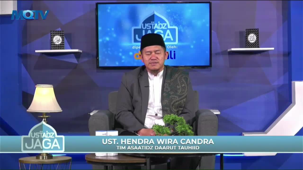 A man in a suit and cap sits in a studio chair, speaking on the Indonesian MQTV program "Ustadz Jaga." The set is simply decorated with a lamp and small plants.
A man in a suit and cap sits in a studio chair, speaking on the Indonesian MQTV program "Ustadz Jaga." The set is simply decorated with a lamp and small plants.