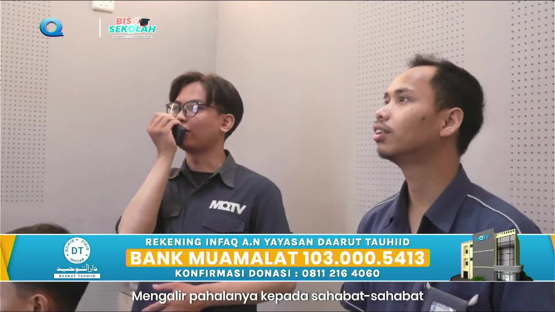 A young man with glasses speaks into a walkie-talkie, his MQTV shirt visible. Beside him, another man turns his head, looking off to the side with his mouth slightly open.