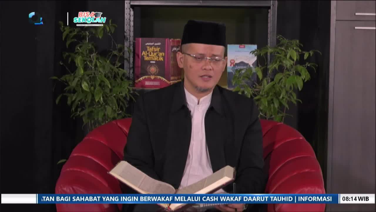 A man in a black peci and jacket sits on a red chair, reading from an open book. Behind him, shelves hold religious texts and a lush green plant.