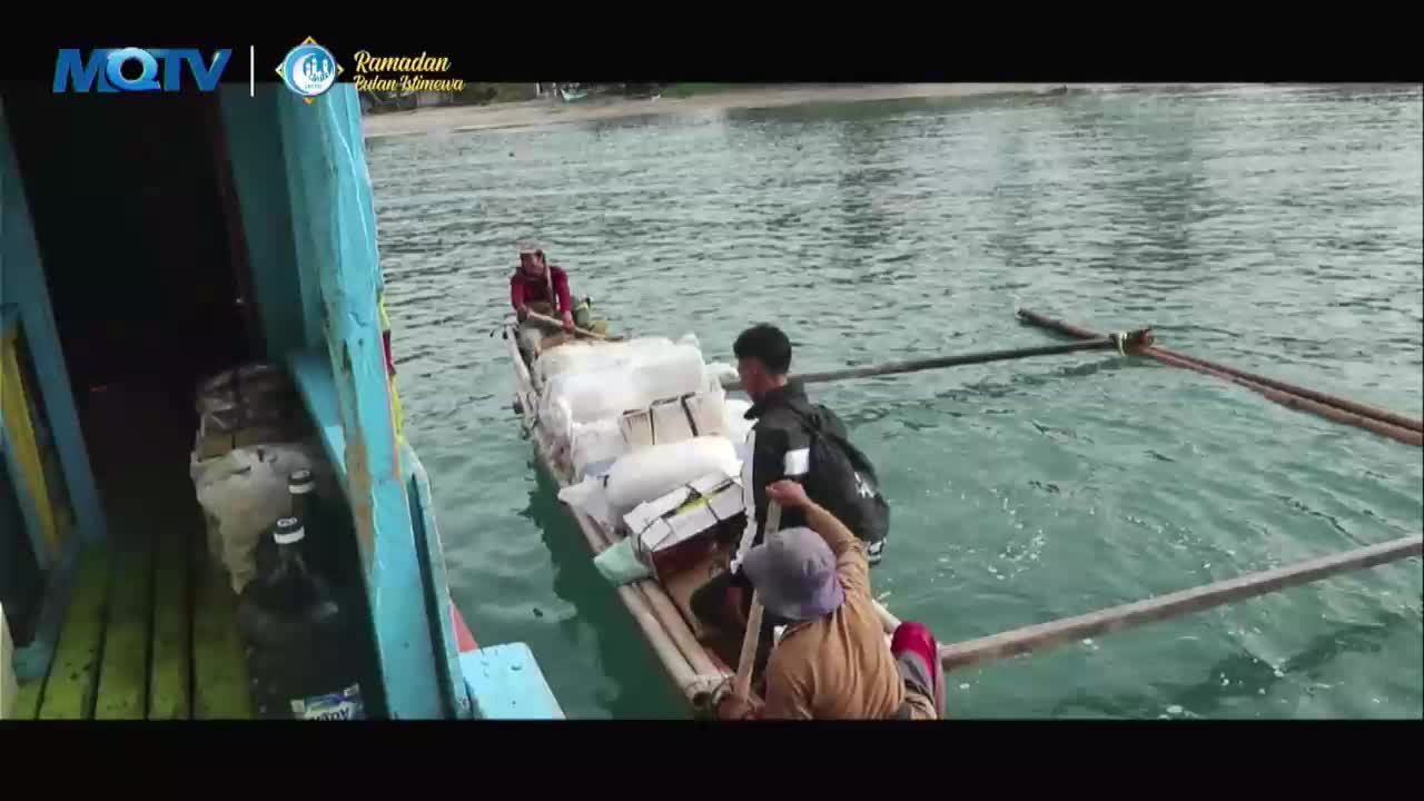 A small boat, laden with white sacks and boxes, is being maneuvered alongside a larger vessel. Three men are working on the smaller boat, one rowing with a long oar. The water is a clear turquoise, reflecting the bright Indonesian sky.