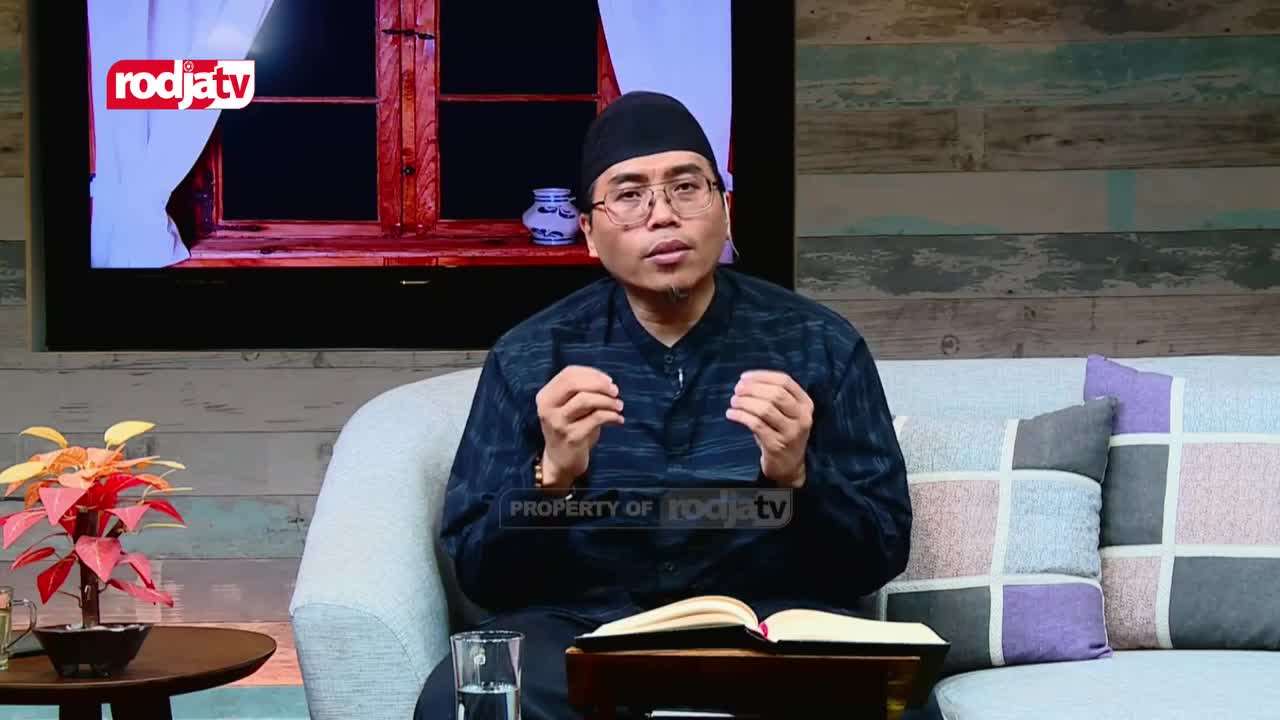 A man in a black peci and patterned shirt speaks with his hands raised, gesturing thoughtfully. A Rodja TV logo is visible on the screen behind him, and an open book rests on a table in front of him. A man in a black peci and patterned shirt speaks with his hands raised, gesturing thoughtfully. A Rodja TV logo is visible on the screen behind him, and an open book rests on a table in front of him.