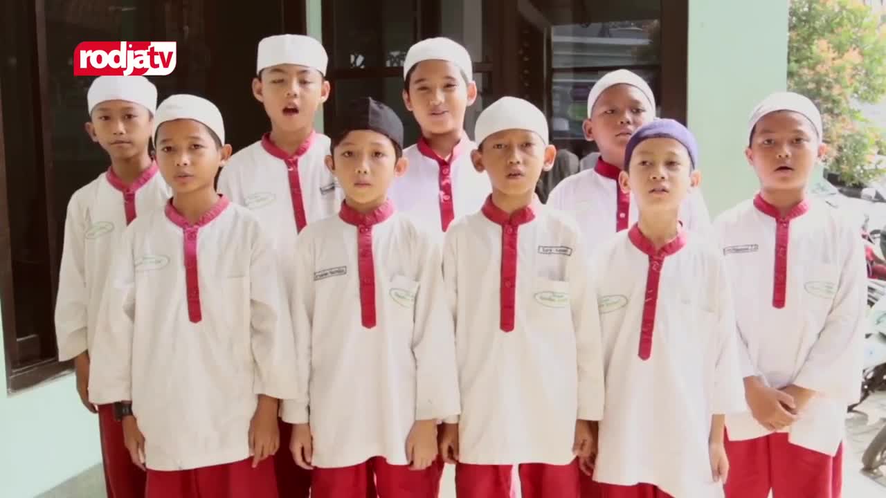 A group of boys in white tunics and red pants stand in front of a building, their mouths open as if singing. The Rodja TV logo appears in the upper left corner of the frame.