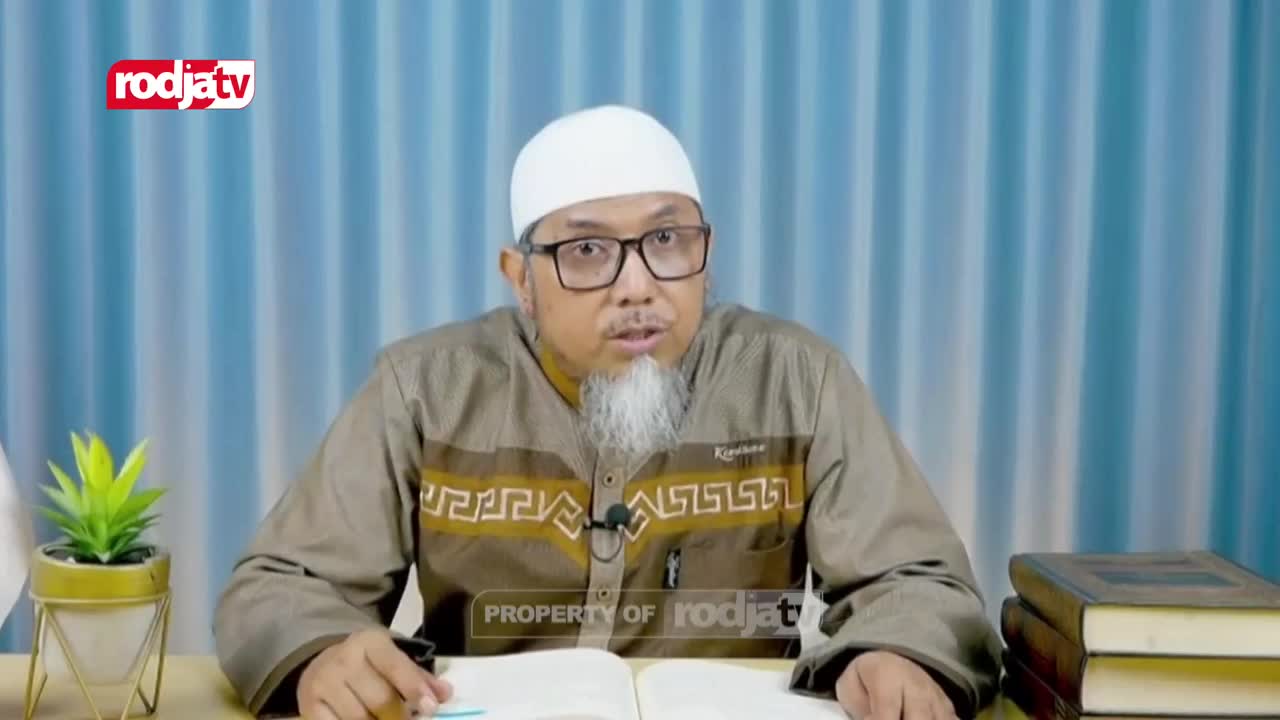 A man with a white cap and glasses speaks, gesturing with his right hand. He wears a brown shirt with a patterned design, and a microphone is clipped to its front.