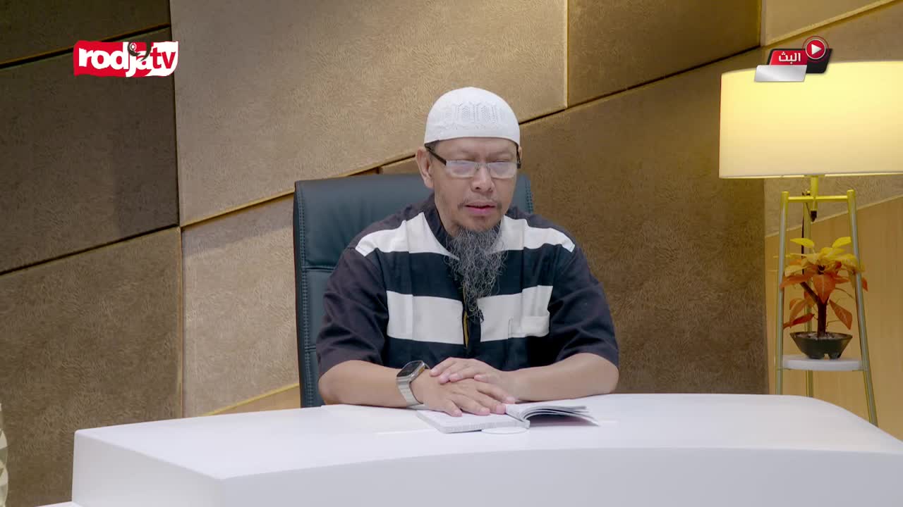 A man in a white peci and a striped shirt sits at a white desk, reading from a book. The Rodja TV logo appears in the upper left corner.