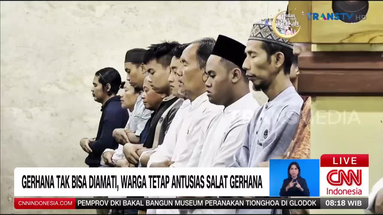 A line of men, some wearing peci caps, stand with their eyes closed, hands clasped. They are participating in a prayer, likely the special prayer for an eclipse, as indicated by the Indonesian text on the screen.