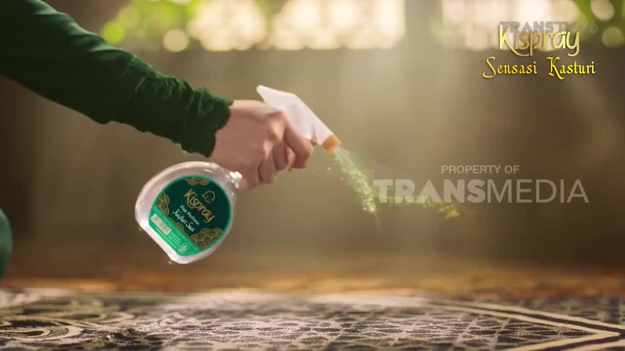 A hand squeezes a spray bottle, releasing a fine mist of green liquid. The mist catches the light as it drifts across the patterned carpet.