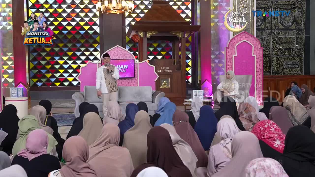 A man in a white jacket and patterned vest speaks into a microphone while standing on a stage. A woman in a white hijab sits on a pink throne to his right, facing a seated audience. The Trans TV logo is visible on a screen behind them.