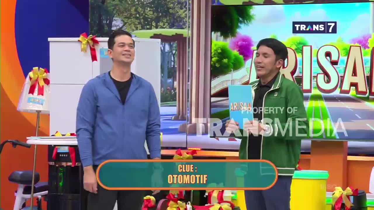 Two men stand on a colorful studio set, one in a blue jacket, the other in green. A large screen behind them displays the Trans 7 logo and the word "ARISAN."