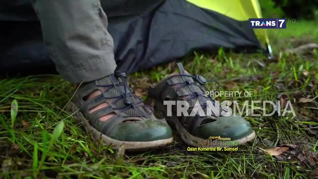 A pair of worn hiking sandals rests on the damp, green grass near a tent. The Trans7 logo is visible in the background, suggesting this is a scene from an Indonesian broadcast.