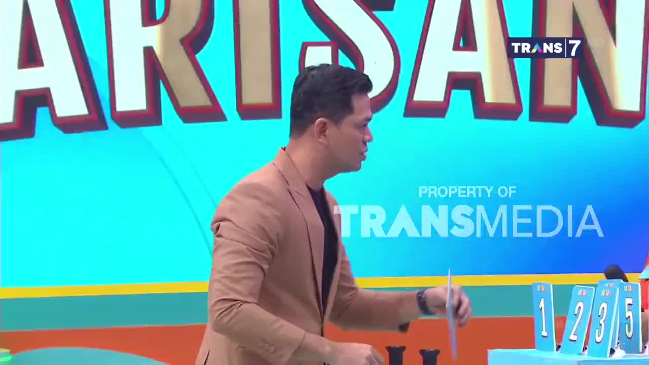 A man in a tan jacket gestures with a card as bright blue number tiles stand before him. The Trans 7 logo is visible behind him. A man in a tan jacket gestures with a card as bright blue number tiles stand before him. The Trans 7 logo is visible behind him.