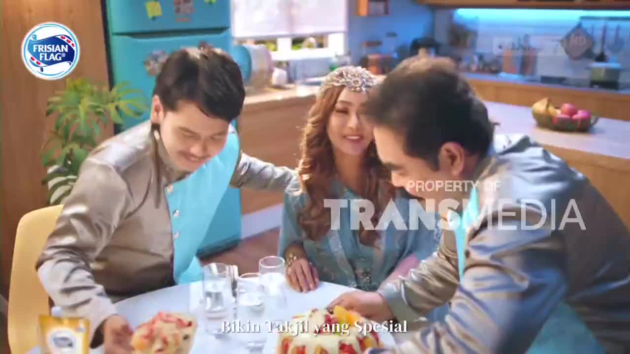 A man in a silver and blue outfit leans forward, reaching for a bowl of fruit salad. Beside him, a woman smiles, her hair adorned with a jeweled headband. Another man, also in a similar outfit, is seated to her left, looking down at the table.