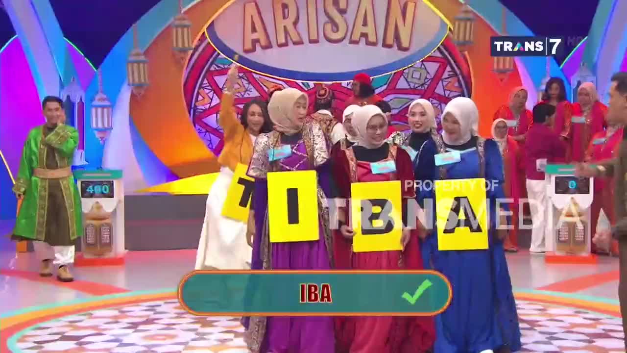 A group of women stand holding large yellow cards with letters on them, forming the word "IBA." The Trans7 logo is visible in the background.