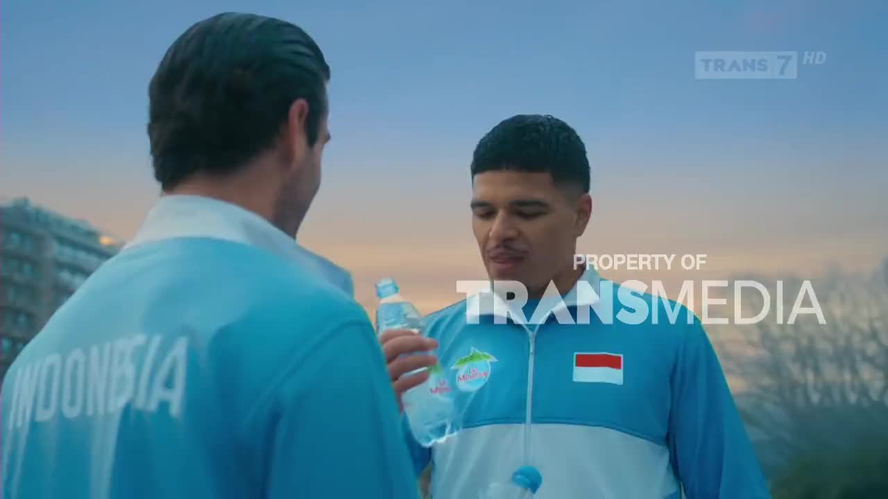 Two men in light blue tracksuits stand facing each other, one holding a bottle of water. The Indonesian flag is visible on the chest of the man on the right.