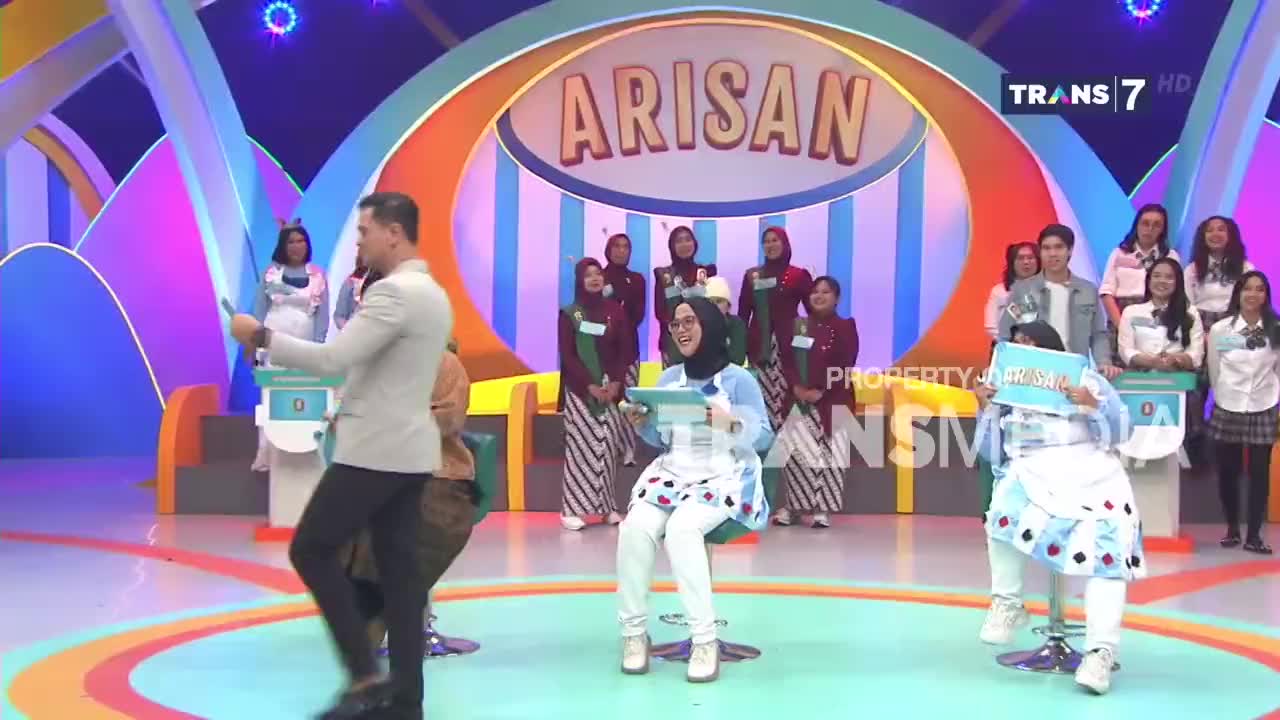 A man in a light suit walks across the stage, holding a tablet. Two women sit on stools, each holding a blue board with text.