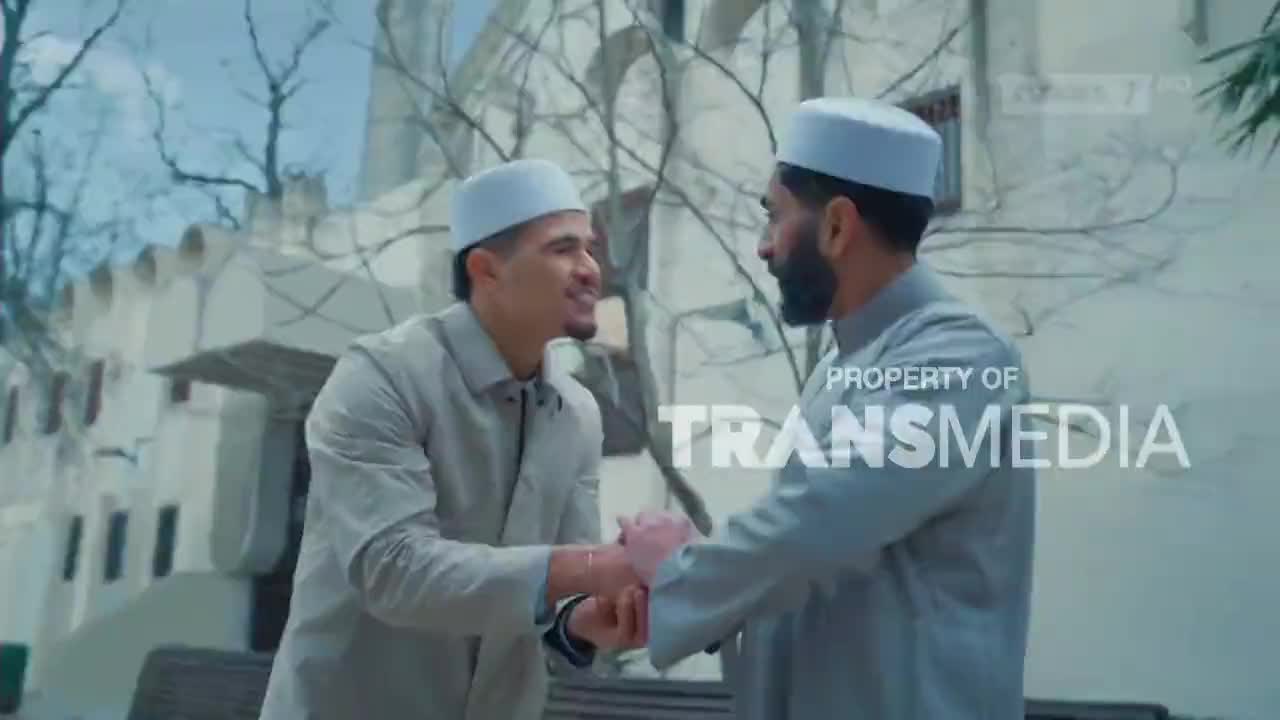 Two men in peci caps shake hands, their smiles friendly against the pale stone of a building. A Trans7 watermark is visible on the right.