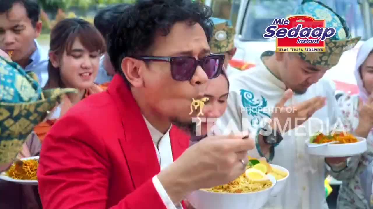 A man in a bright red jacket slurps noodles from a bowl, his sunglasses reflecting the scene. Beside him, others in traditional Indonesian attire also enjoy their Mie Sedaap instant noodles. A man in a bright red jacket slurps noodles from a bowl, his sunglasses reflecting the scene. Beside him, others in traditional Indonesian attire also enjoy their Mie Sedaap instant noodles.