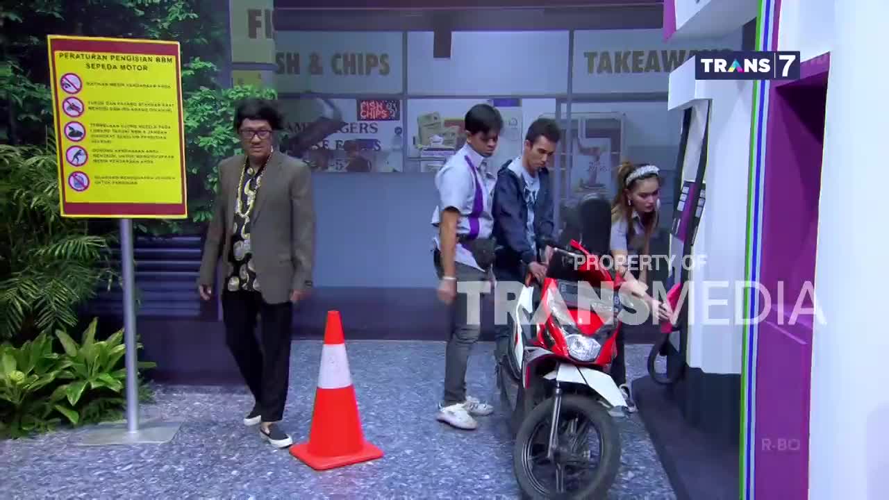 A woman is pumping gas into a red scooter at a station, while a man in a blue jacket stands beside her. A Trans7 logo is visible on the gas pump. A woman is pumping gas into a red scooter at a station, while a man in a blue jacket stands beside her. A Trans7 logo is visible on the gas pump.