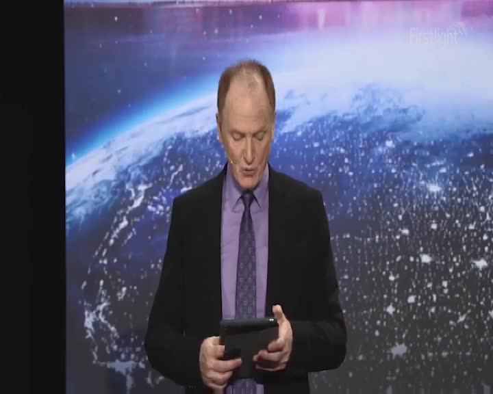 A man in a suit reads from a tablet, his gaze fixed downwards. Behind him, a backdrop of a nighttime Earth, with the "Firstlight" logo visible in the upper right corner, is lit up.
