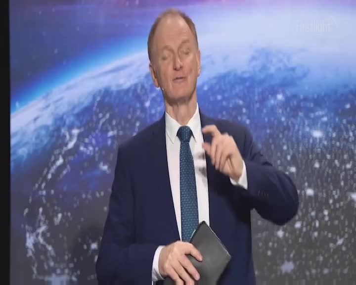 A man in a suit gestures with his right hand, holding a dark book in his left. Behind him, a large screen displays a view of Earth from space, with lights twinkling across continents.