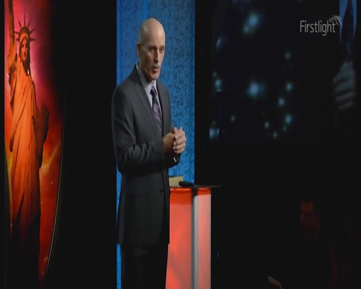 A man in a suit stands at a podium, his hands clasped. Behind him, a large image of the Statue of Liberty glows with an orange light. A man in a suit stands at a podium, his hands clasped. Behind him, a large image of the Statue of Liberty glows with an orange light.