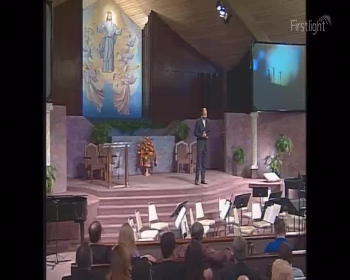 A man in a suit stands on a raised platform, holding a book. Behind him, a large mural depicts Jesus with angels, and a screen displays a blue, abstract image. A man in a suit stands on a raised platform, holding a book. Behind him, a large mural depicts Jesus with angels, and a screen displays a blue, abstract image.